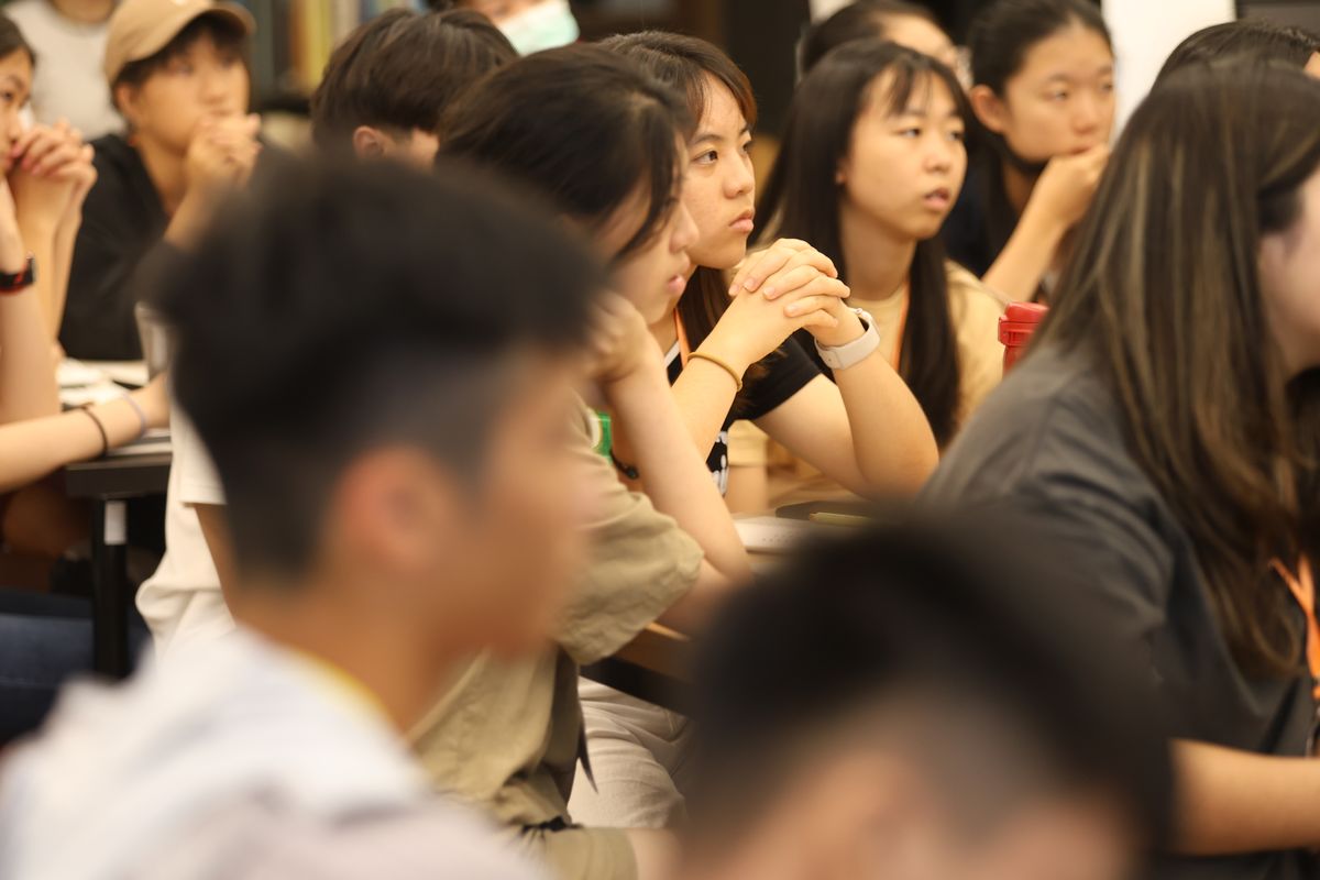 一排高中生坐著聽台上的人講話，前景是兩個模糊的後腦勺